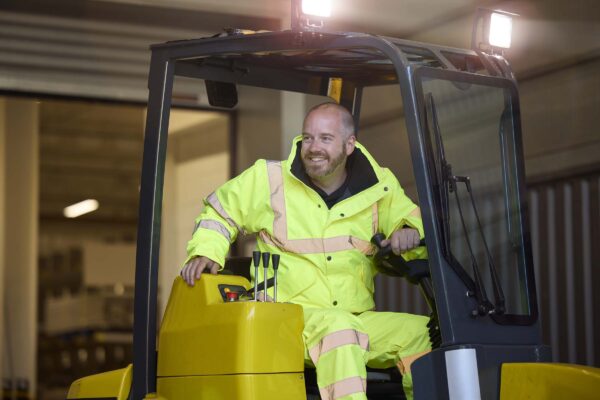 “Forklift driver wearing high-visibility yellow workwear, operating machinery comfortably in a warehouse environment.”